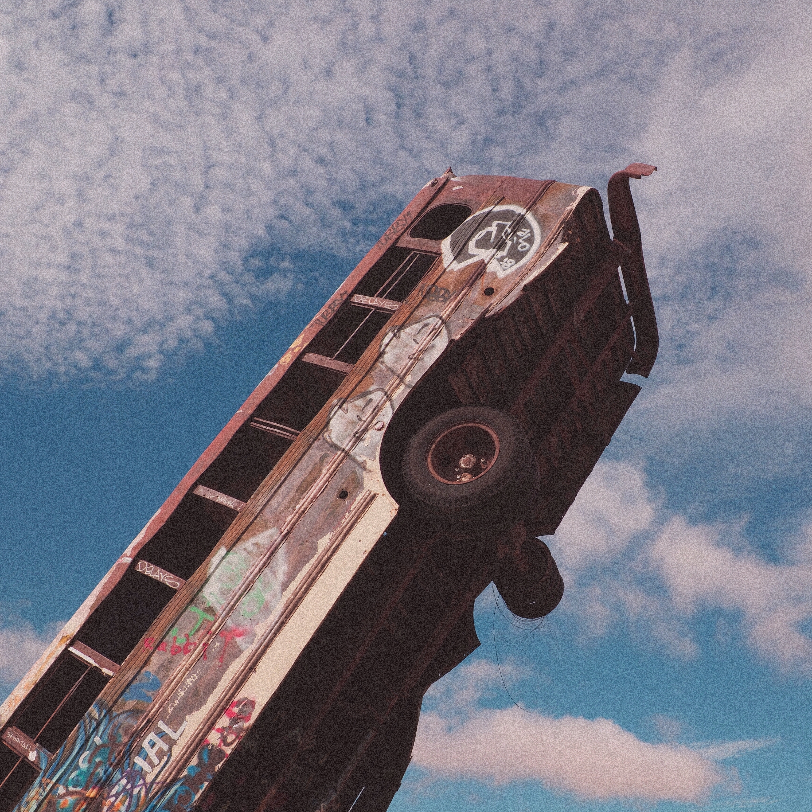 remains of a rust and graffiti covered bus, sticking straight up in the air in front of a blue cloudy sky
