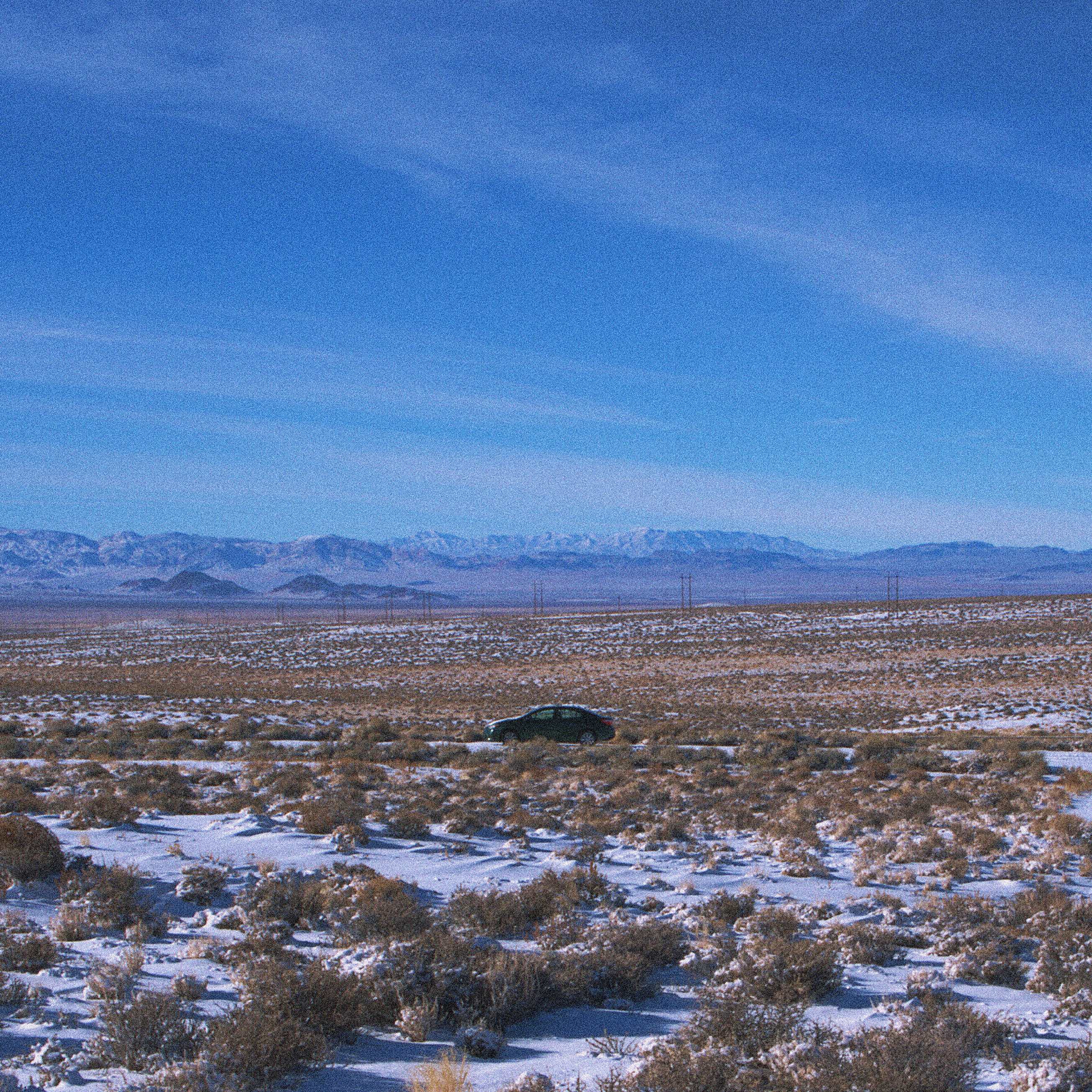 Far away view of a green toyota corolla parked alone in a snowy field full of sage grass with distan mountains in the background