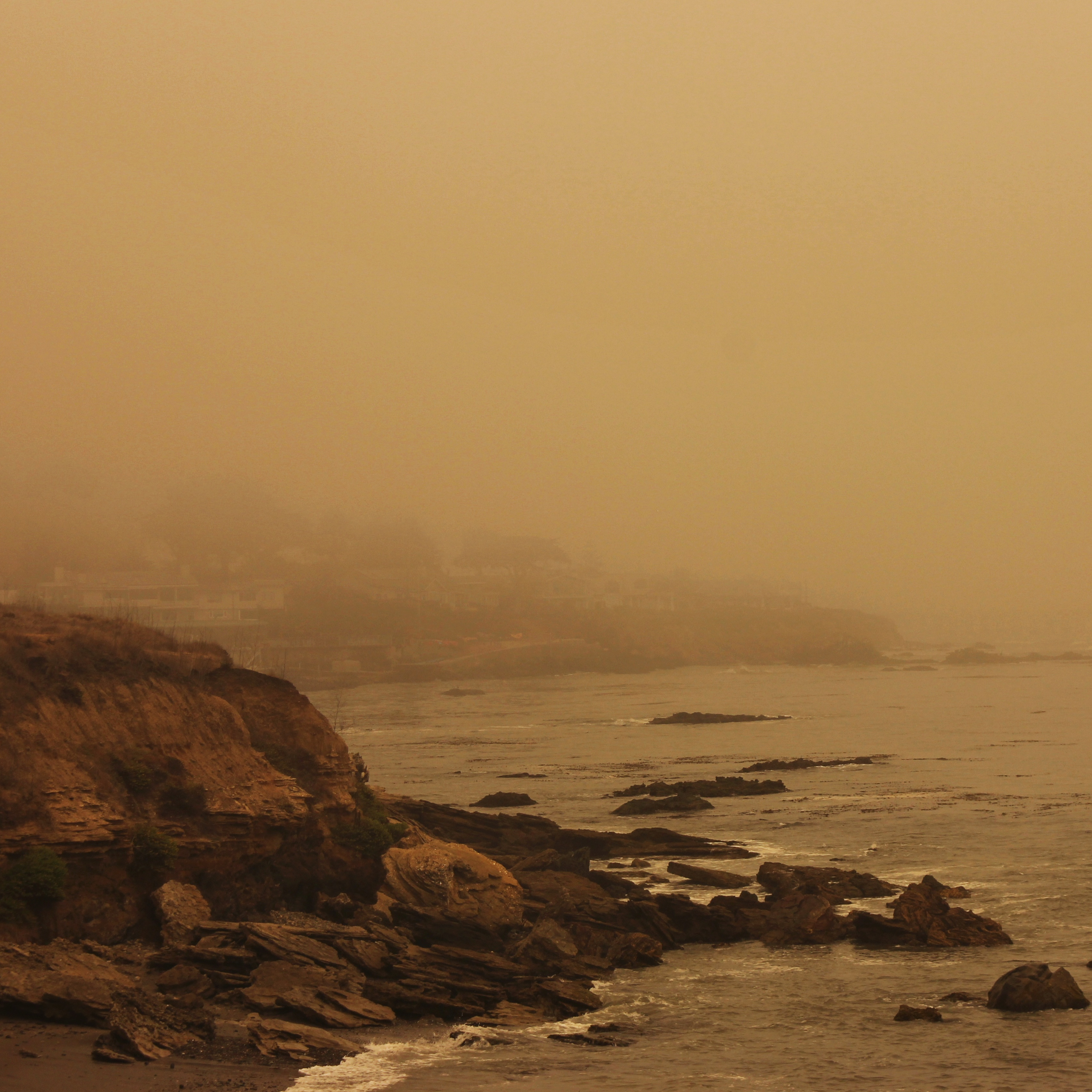 a rocky cliff overlooking the ocean and a town in the background, barely visible behind thick yellow fog