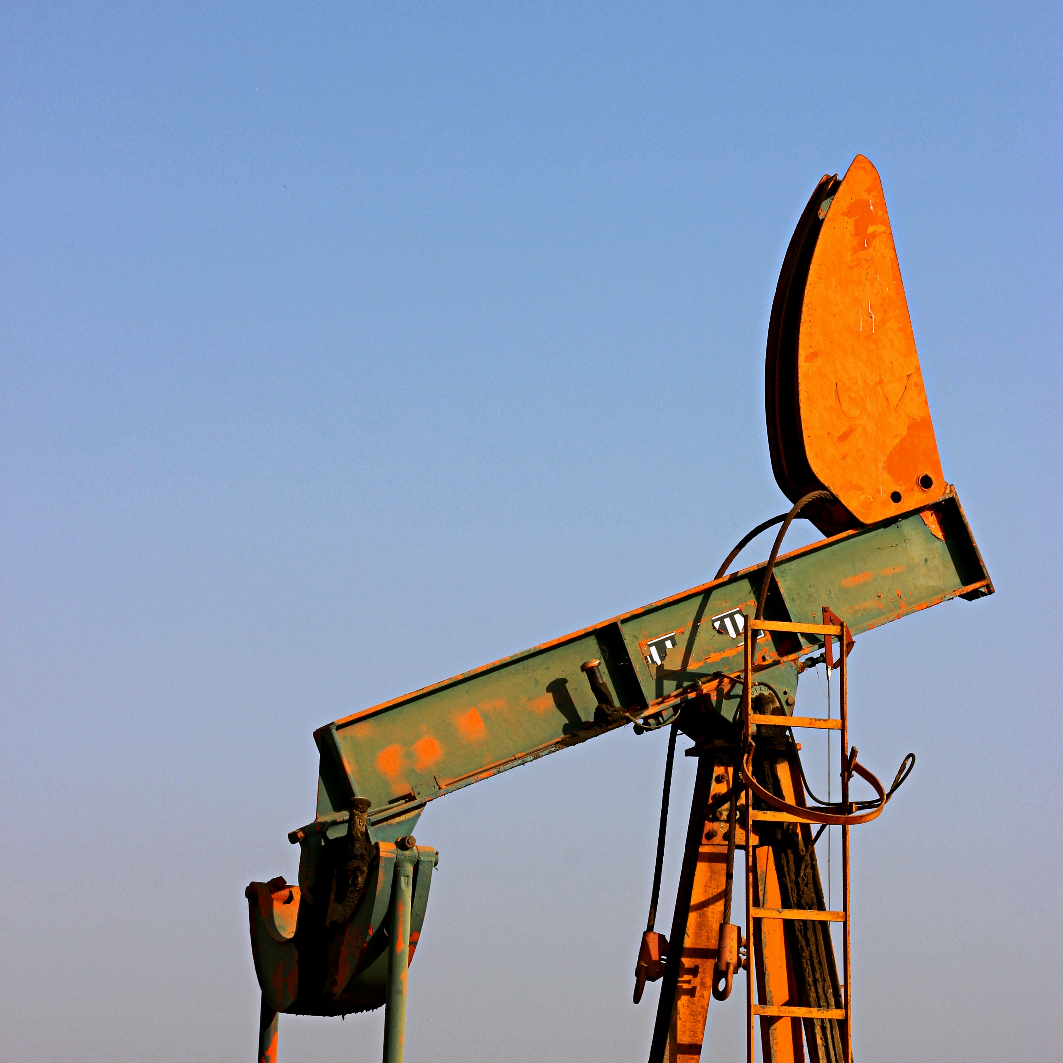 close-up of a rusty orange and green oil drill contrasted against a blank blue sky