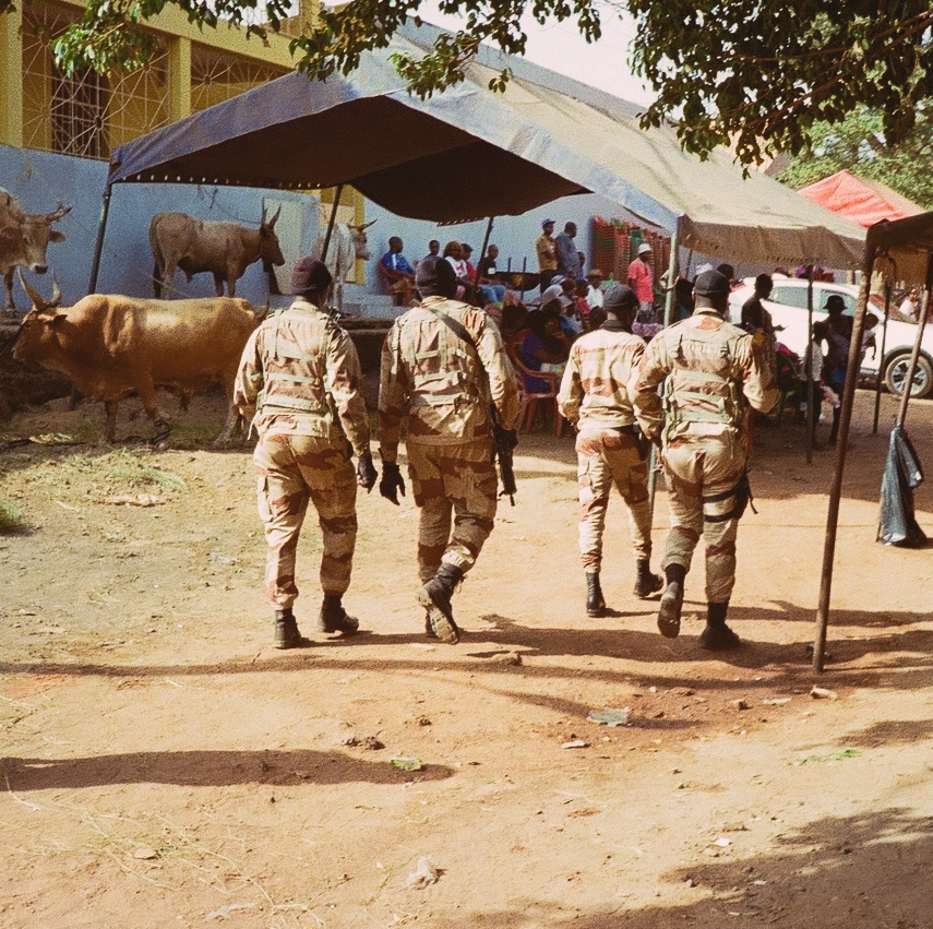 four soldiers walking up a dirt street in Bissau with bulls and a gathered crowd in the background